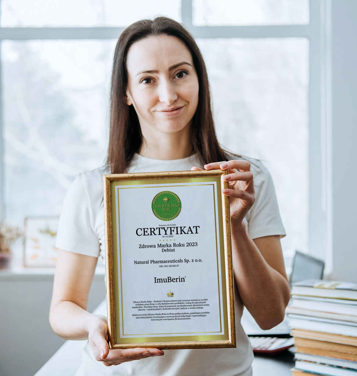 Natural Pharmaceuticals ImuBerin® Kobieta pokazująca certyfikat Zdrowa Marka Roku 2023 dla firmy Natural Pharmaceuticals i produktu ImuBerin®.