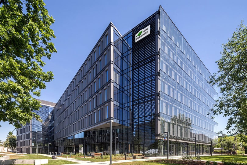 Natural Pharmaceuticals ImuBerin® Nowoczesny szklany budynek biurowy otoczony zielenią pod błękitnym niebem.